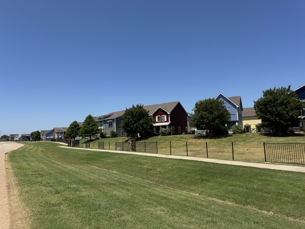 Natural-grass common area beside a sidewalk and fenced residential community