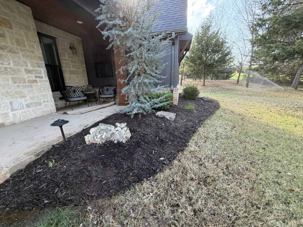 Dark mulch surrounding landscape plants and decorative stones beside a front porch