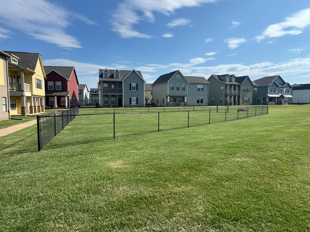 Natural-grass dog park enclosed by black chain-link fencing beside community homes